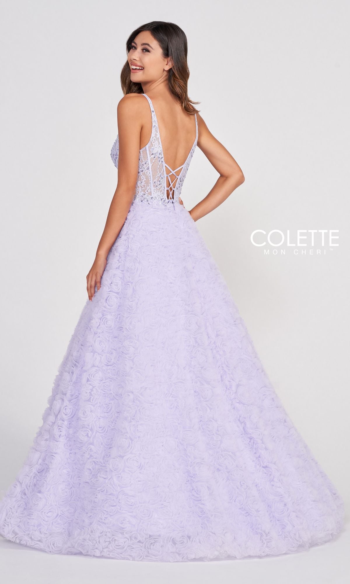 A woman faces away, turning her head to the side, wearing the Colette Sheer-Bodice Rosette Beaded Lace Ball Gown featuring a light purple hue, lace-up back, and floral textured skirt.